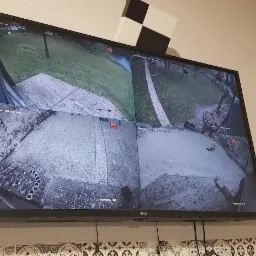 Ekran monitora LCD z widokiem z czterech kamer monitoringu CCTV, prezentujący podwórko z trawnikiem, ścieżką i elementami dekoracyjnymi.