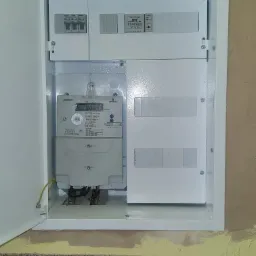 Otwarta biała skrzynka rozdzielcza z licznikiem energii elektrycznej Norax 3 i zabezpieczeniami na ścianie z żółtą farbą, widoczne przewody i uziemienie.