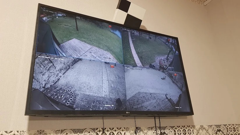 Ekran monitora LCD z widokiem z czterech kamer monitoringu CCTV, prezentujący podwórko z trawnikiem, ścieżką i elementami dekoracyjnymi.
