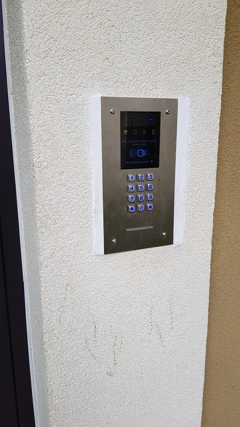 Nowoczesny, metalowy panel domofonu z podświetlanymi przyciskami, zamontowany na teksturowanej, beżowej ścianie budynku.