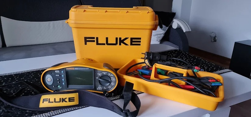 Zestaw żółtych narzędzi pomiarowych Fluke, w tym miernik wielofunkcyjny, przewody i skrzynka, ułożone na stole w domowym otoczeniu.