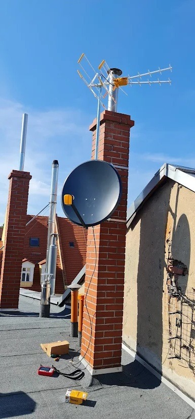 Antena satelitarna i antena naziemna zamontowane na kominie z czerwonej cegły na dachu, widoczne narzędzia i pudełka po akcesoriach.