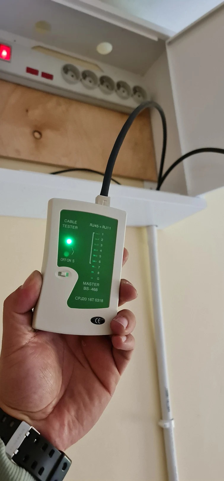 Testowanie kabla sieciowego RJ45 testerem w dłoni, widoczna skrzynka z gniazdkami elektrycznymi w tle.