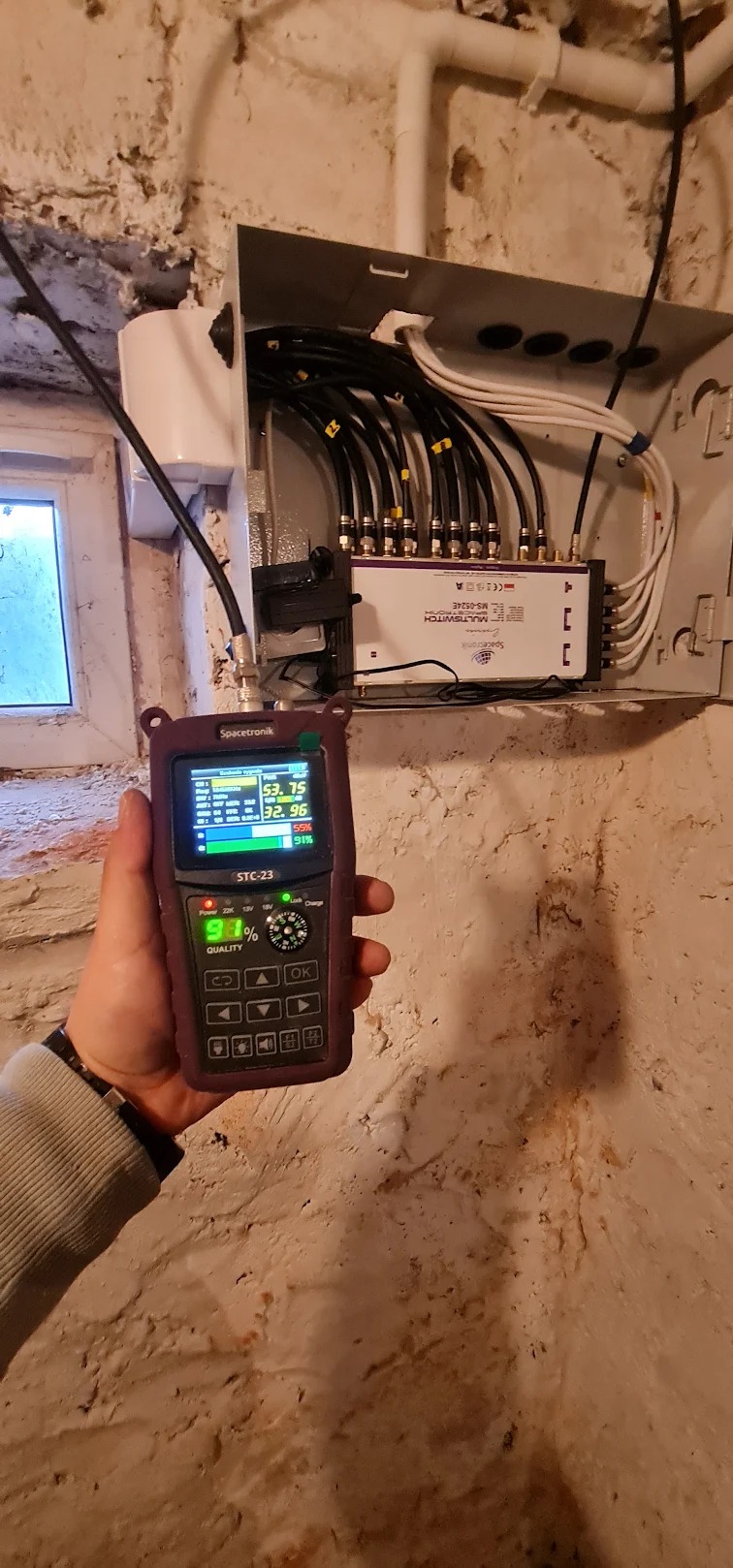 Spacetronik STC-23 mierzy jakość sygnału w instalacji multiswitcha MS-0524E z licznymi kablami koncentrycznymi w szarej skrzynce na tle surowej, pomalowanej ściany w piwnicy.