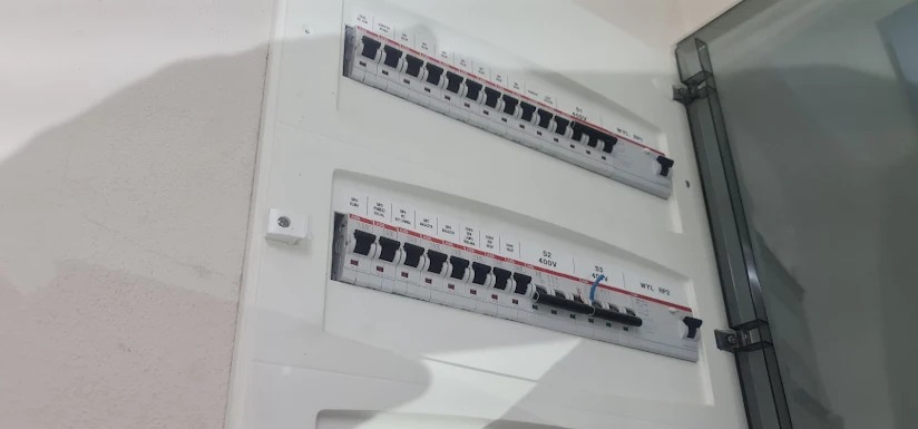Dwa rzędy wyłączników nadprądowych w białej skrzynce rozdzielczej, jeden wyłącznik w pozycji wyłączonej, widoczna ściana i fragment przezroczystych drzwi skrzynki.