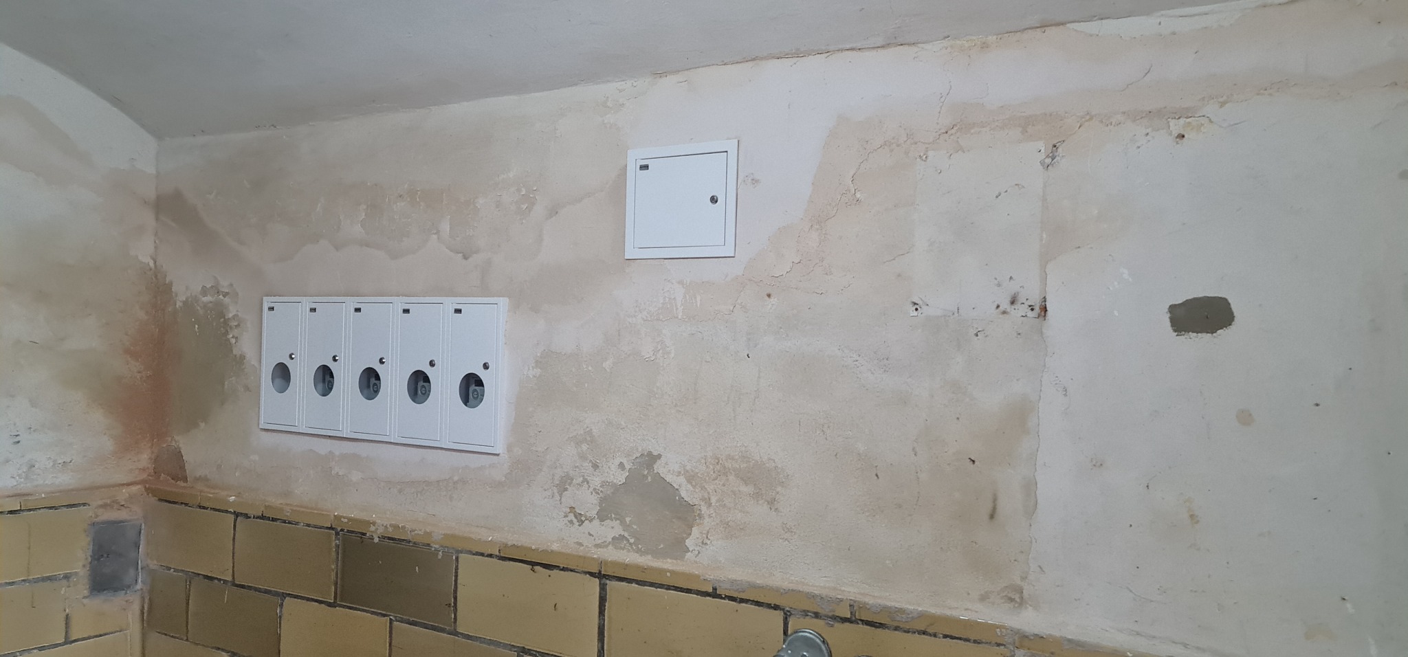 Białe skrzynki licznikowe z okrągłymi okienkami na tle ściany w stanie surowym, z widocznymi śladami wilgoci i ubytkami tynku, oraz fragmentem ceramicznej okładziny.