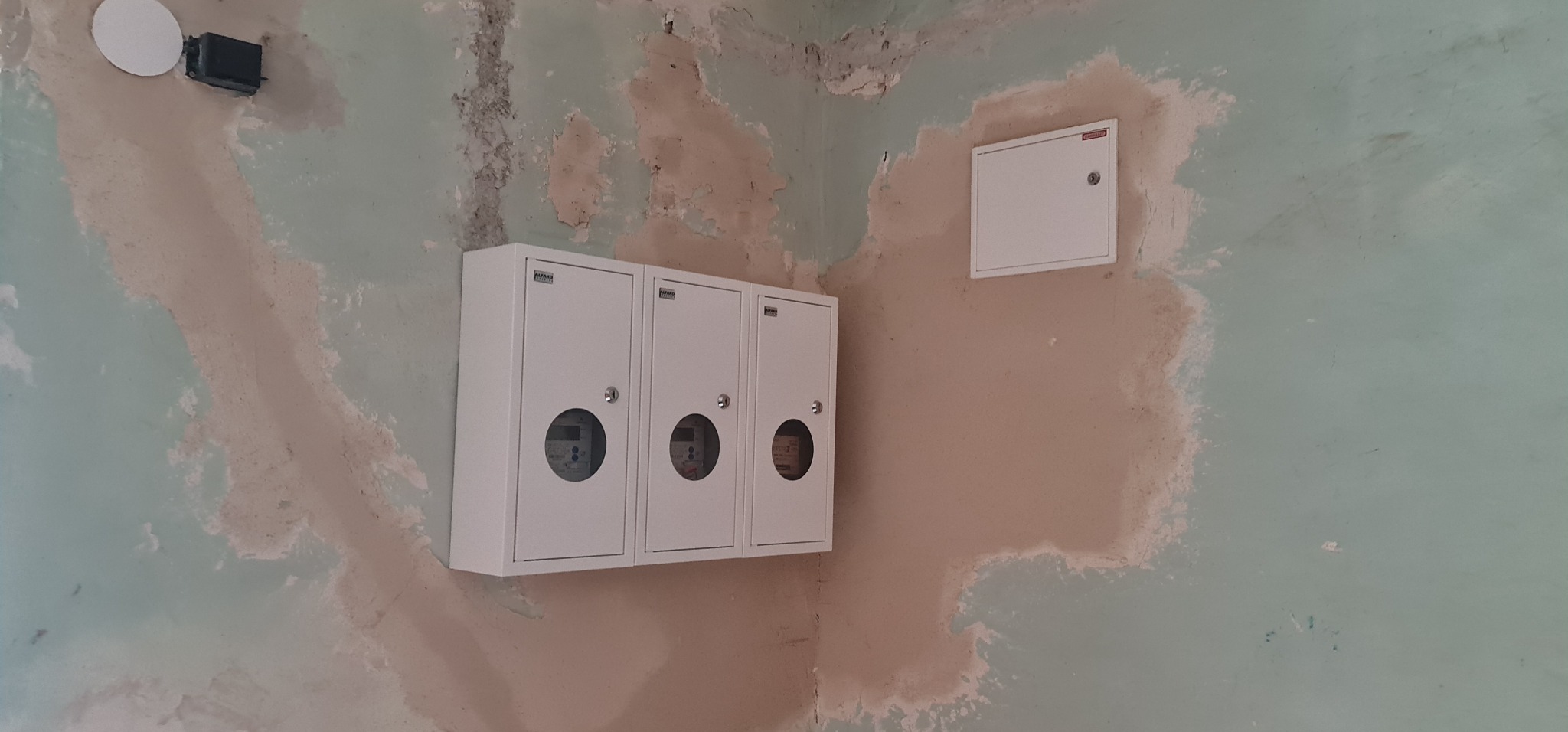 Trzy białe skrzynki z licznikami na ścianie w trakcie remontu, widoczne ślady starej farby i szpachli.