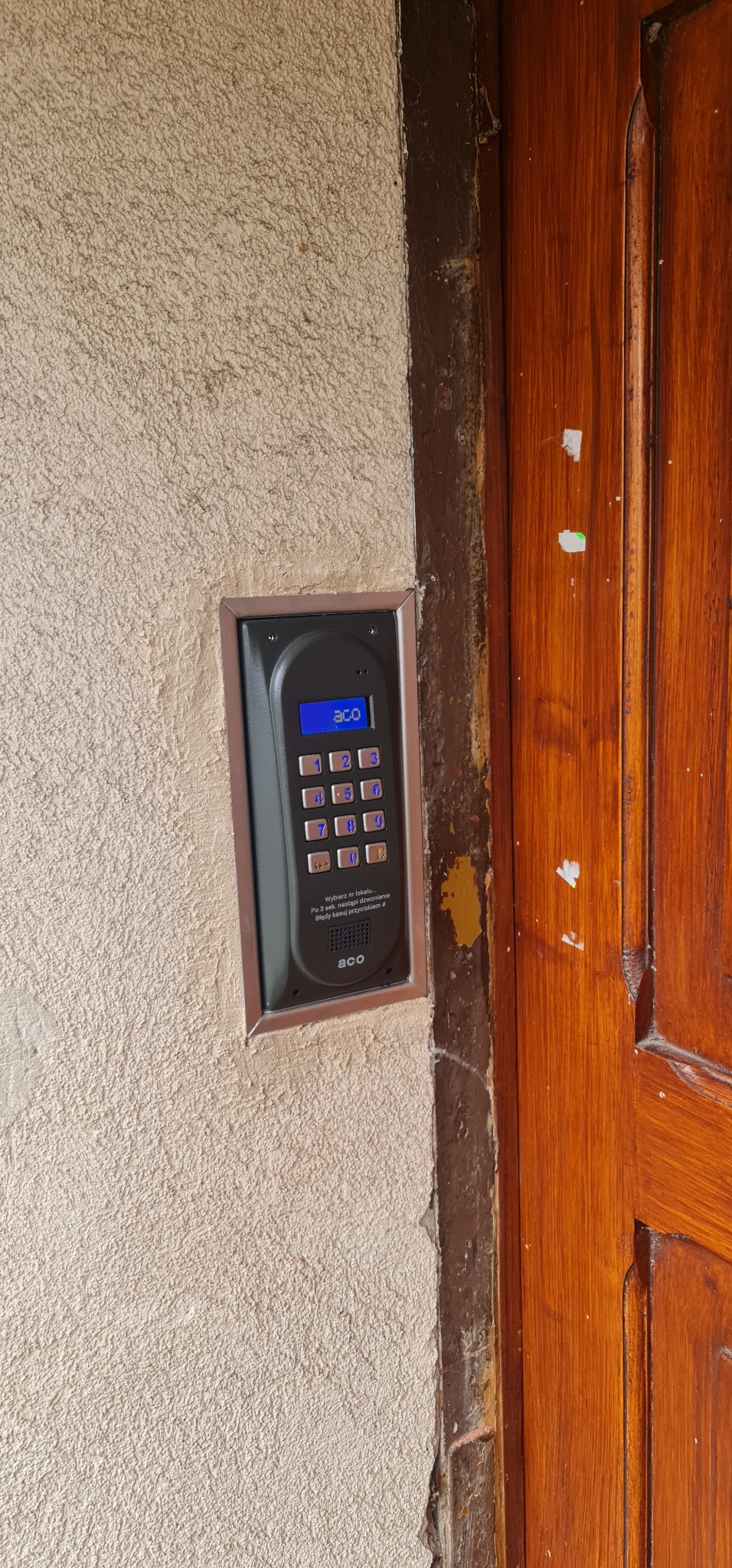 Zamontowany, czarny domofon cyfrowy ACO z niebieskim wyświetlaczem i klawiaturą numeryczną, wmurowany w szorstką ścianę obok drewnianych drzwi z widocznymi śladami użytkowania.