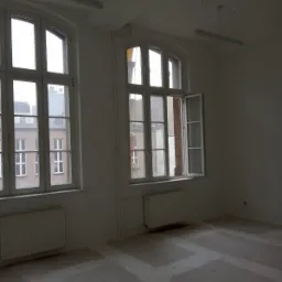 Surowe wnętrze pokoju z dwoma oknami, grzejnikami i przygotowanym podłożem pod podłogę, widok na budynki za oknem.