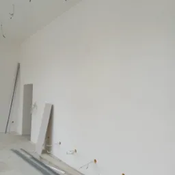 Wnętrze w trakcie remontu: widoczne profile metalowe, płyta gipsowa oparta o ścianę, wystające kable elektryczne ze ściany i sufitu, surowy beton na podłodze.