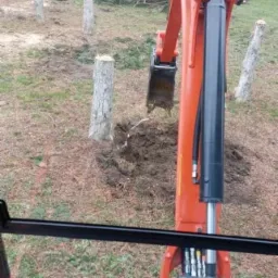 Pomarańczowa mini-koparka Kubota z łyżką kopie dół w ziemi, widok z kabiny operatora, w tle widoczne ścięte pnie drzew i budynek gospodarczy.