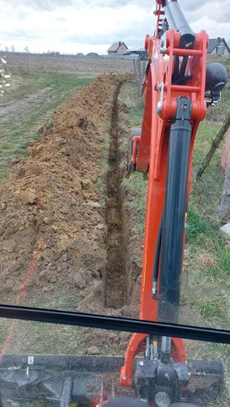 Wąski wykop pod instalacje wykonany pomarańczową mini koparką Kubota na polu z widocznym domem w tle, widok z kabiny operatora przez szybę.