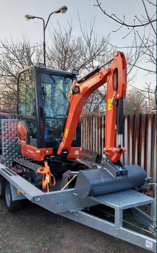 Pomarańczowa minikoparka Kubota zabezpieczona pasami transportowymi na metalowej przyczepie, widoczne oświetlenie uliczne w tle.