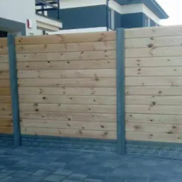 Drewniana konstrukcja ogrodzenia z poziomych desek, wsparta na betonowych słupkach, z widocznym fragmentem nawierzchni z kostki brukowej i elewacji budynku w tle.