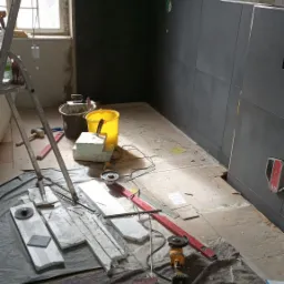 Montaż ciemnych płytek w łazience w trakcie remontu, widoczne narzędzia glazurnicze, poziomica, szlifierka kątowa, drabina i przygotowane płytki.