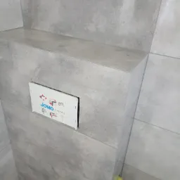 Szara, wykończona ściana łazienki z zamontowanym stelażem podtynkowym WC marki JOMO, widoczne otwory na rury i żółty element montażowy.