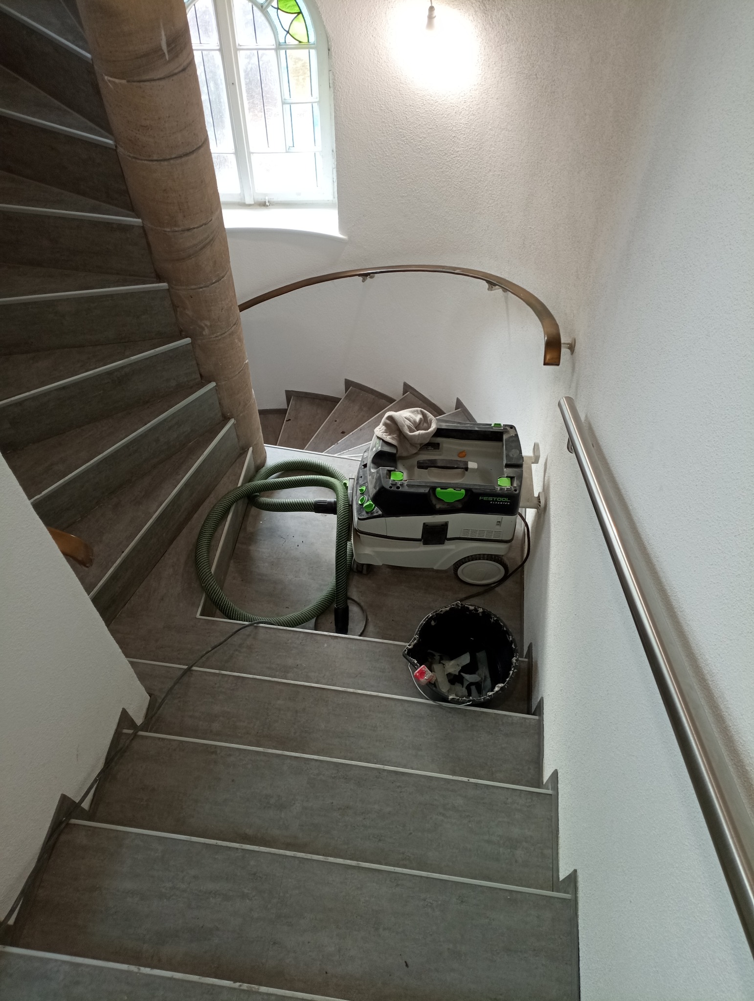 Widok z góry na spiralne schody z szarymi stopniami, na podeście stoi odkurzacz przemysłowy Festool z zielonym wężem oraz czarne wiadro z odpadami, w tle okno z witrażem.
