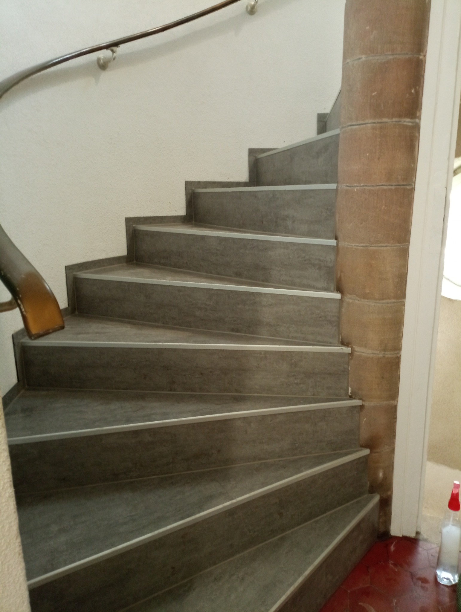 Betonowe schody spiralne z metalowymi krawędziami zabezpieczającymi, widoczne z dołu, z fragmentem brązowej poręczy i kolumny obłożonej kamieniem.