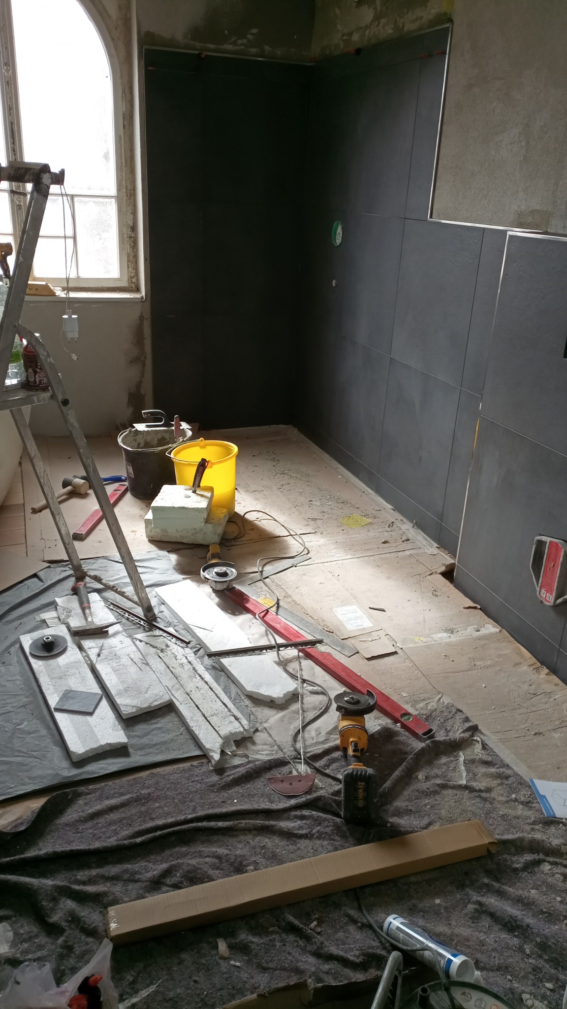 Montaż ciemnych płytek w łazience w trakcie remontu, widoczne narzędzia glazurnicze, poziomica, szlifierka kątowa, drabina i przygotowane płytki.