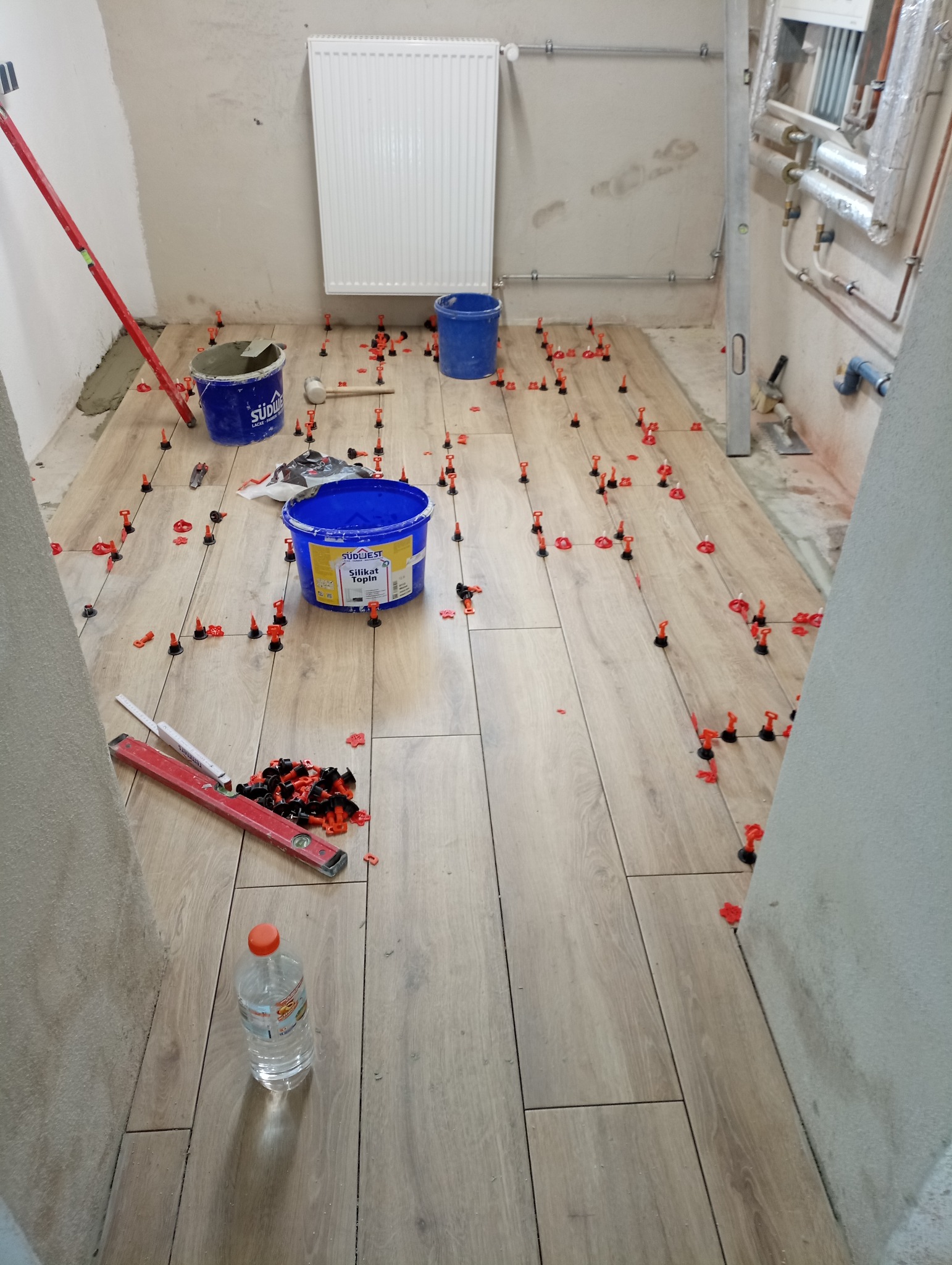 Podłoga w trakcie układania płytek imitujących drewno, widoczne poziomice, wiadra z zaprawą, system poziomowania płytek oraz butelka wody.