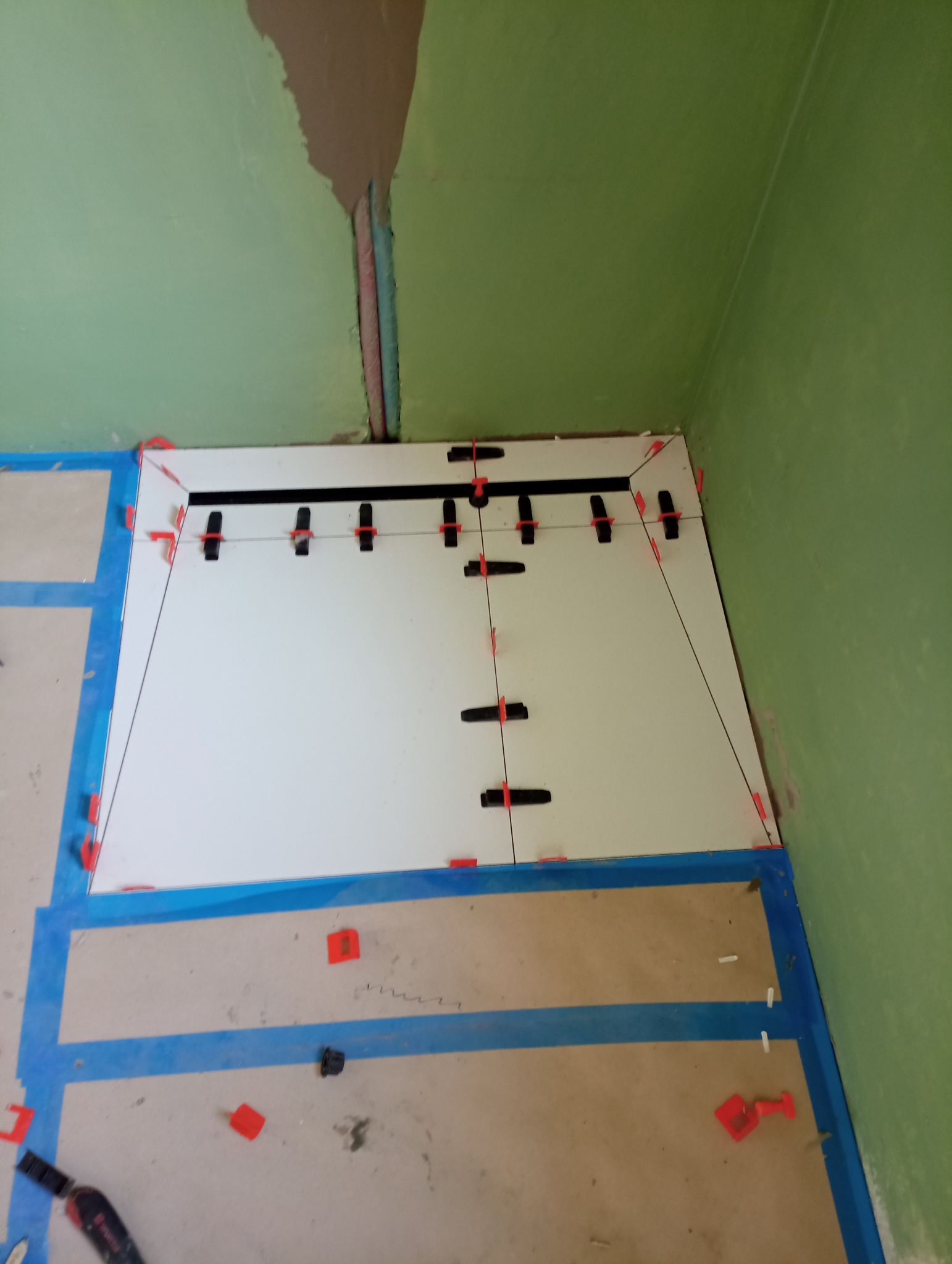 Układanie białych płytek podłogowych z użyciem poziomowania krzyżykami w pomieszczeniu z zielonymi ścianami i widocznymi rurami w ścianie, zabezpieczonymi niebieską taśmą.