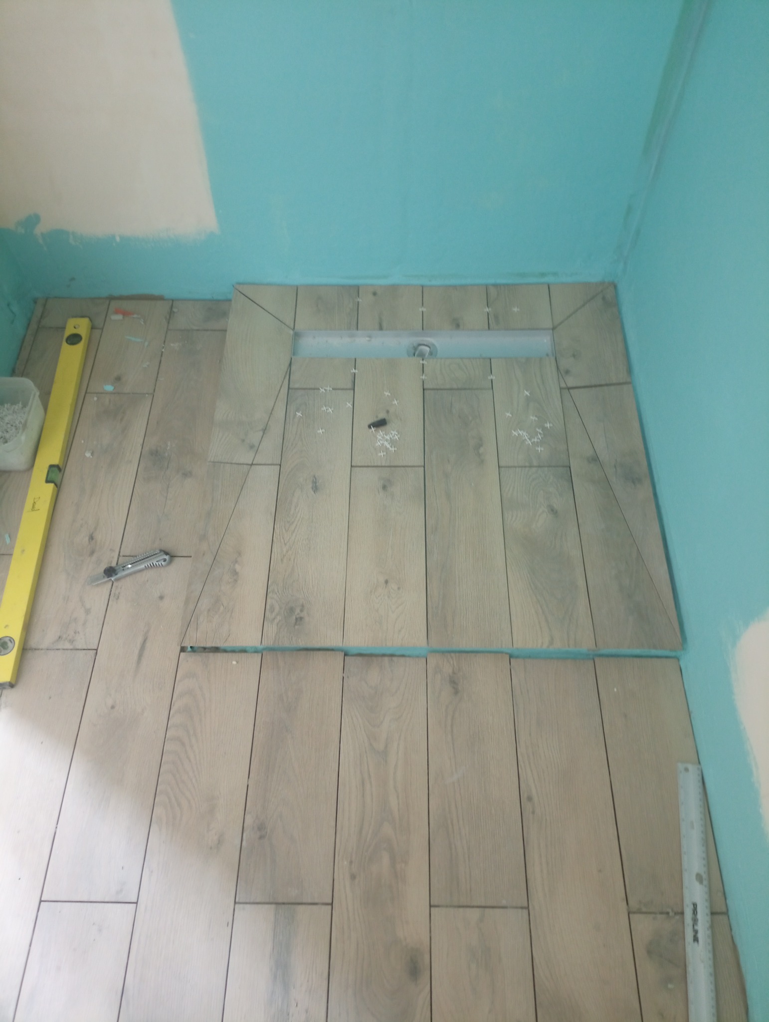 Układanie płytek imitujących drewno w strefie prysznicowej z odpływem liniowym; widoczny poziomica, nóż do cięcia, krzyżyki dystansowe i niepomalowane fragmenty ścian.