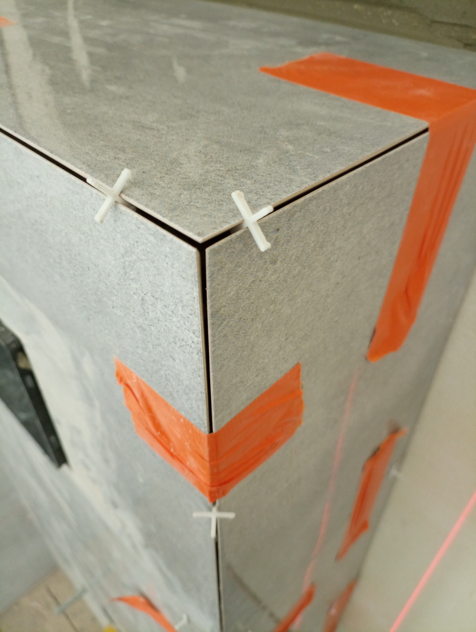 Fragment ściany wyłożonej szarymi płytkami ceramicznymi, zabezpieczonymi pomarańczową taśmą i krzyżykami dystansowymi, widoczna wiązka lasera wyznaczająca poziom.