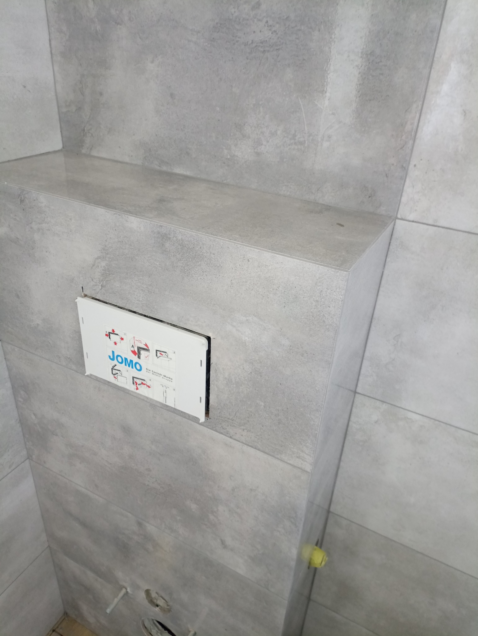 Szara, wykończona ściana łazienki z zamontowanym stelażem podtynkowym WC marki JOMO, widoczne otwory na rury i żółty element montażowy.