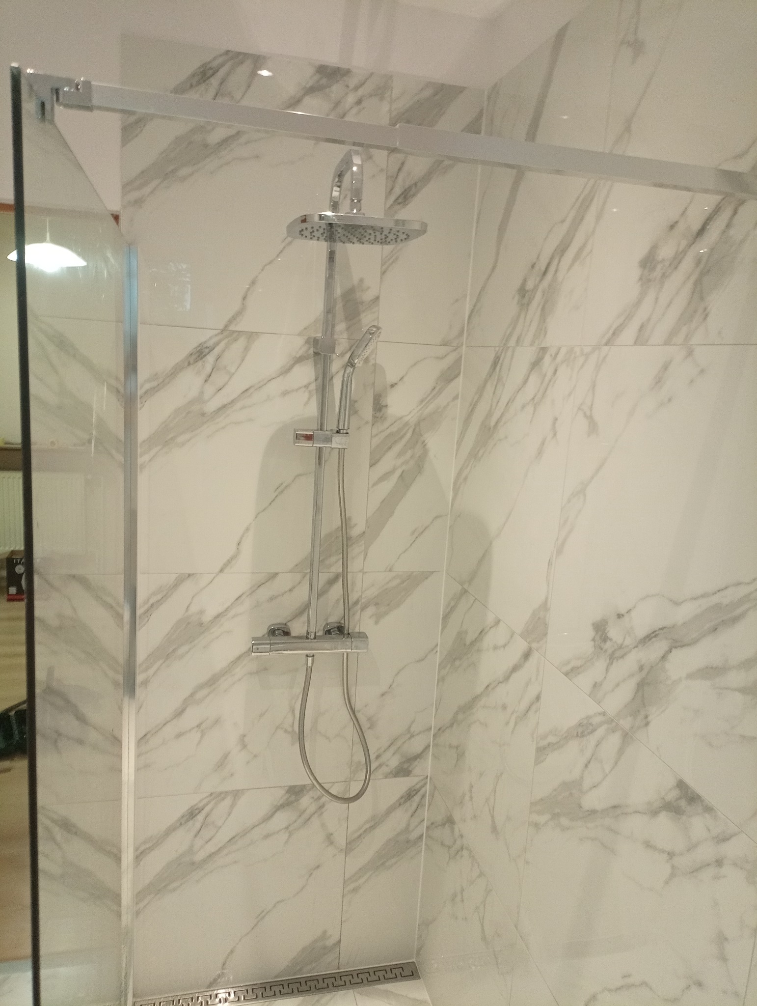 Kabina prysznicowa z deszczownicą i baterią termostatyczną, wykończona płytkami imitującymi marmur z dekoracyjnym odpływem liniowym.