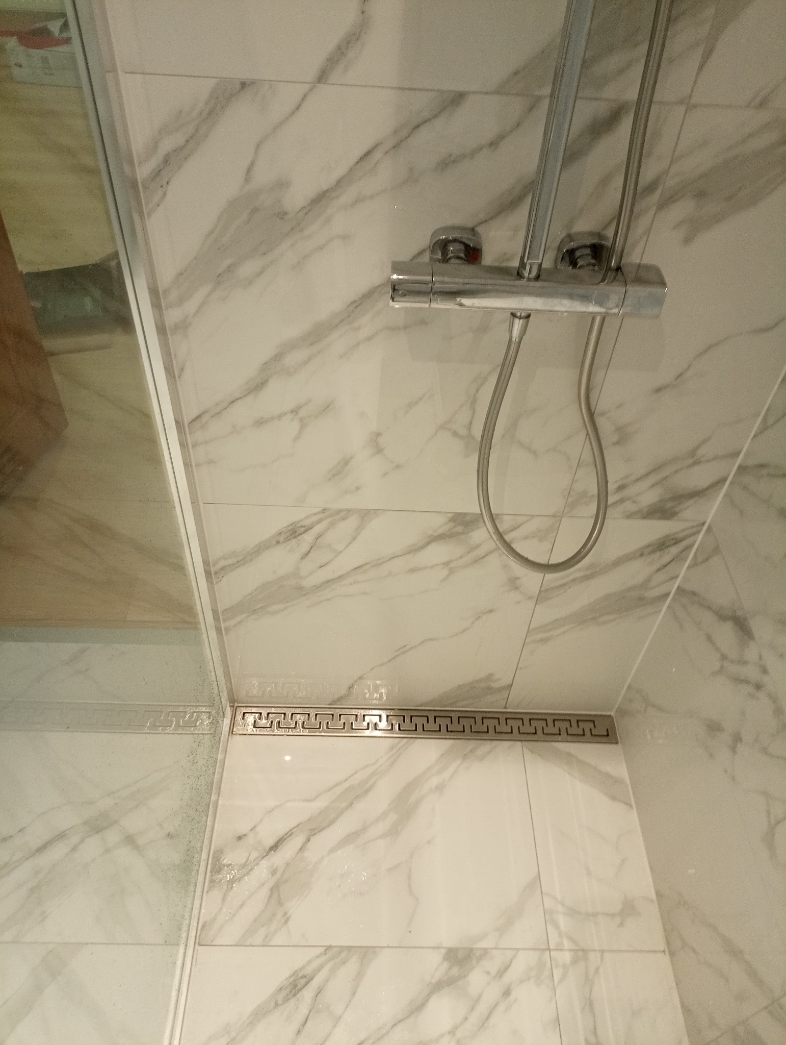 Białe marmurowe płytki w kabinie prysznicowej z odpływem liniowym ozdobionym greckim wzorem i chromowaną baterią.