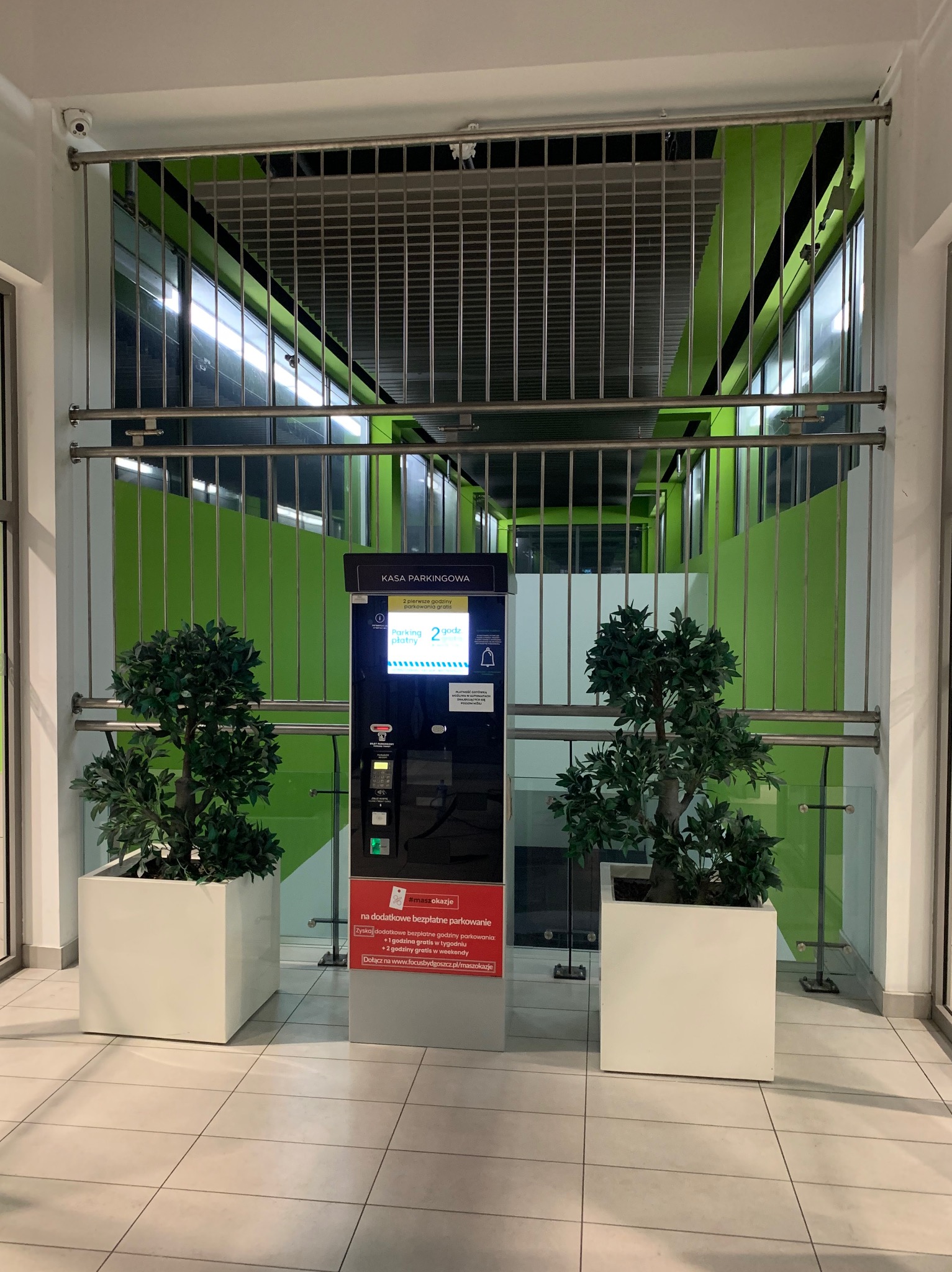 Metalowa krata zabezpieczająca w tle kasy parkingowej, flankowana przez dwa sztuczne drzewka w białych donicach, na tle zielonych ścian budynku.