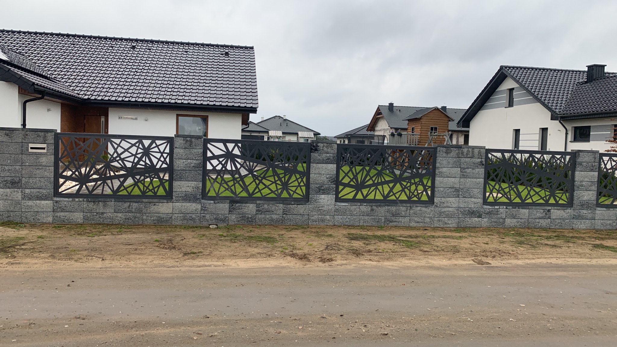 Nowoczesne przęsła ogrodzeniowe z geometrycznym wzorem, osadzone w betonowym murze, widok na domy jednorodzinne w tle.