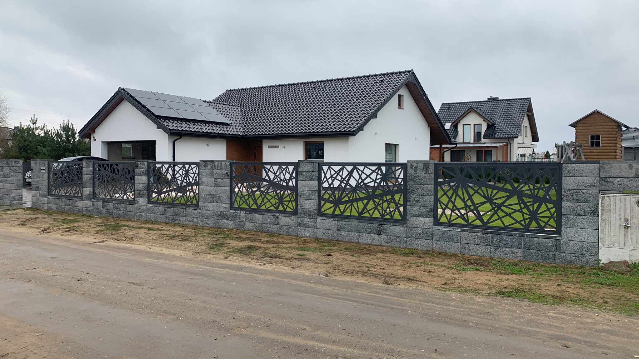 Nowoczesna, ażurowa, metalowa konstrukcja ogrodzeniowa z szarej kostki betonowej przed domem z panelami słonecznymi na dachu w pochmurny dzień.
