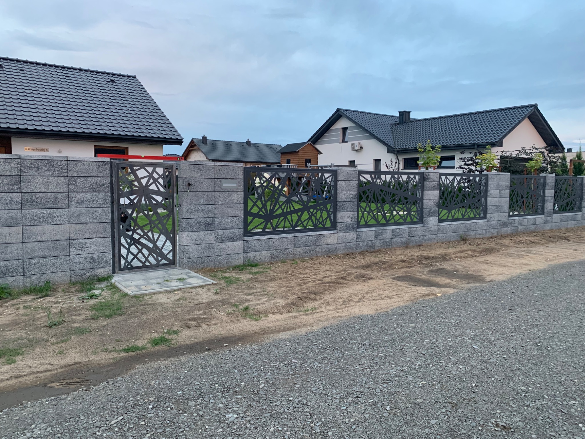 Nowoczesna brama wjazdowa i ogrodzenie z szarego betonu architektonicznego i metalowych paneli o geometrycznym wzorze na tle domów jednorodzinnych.