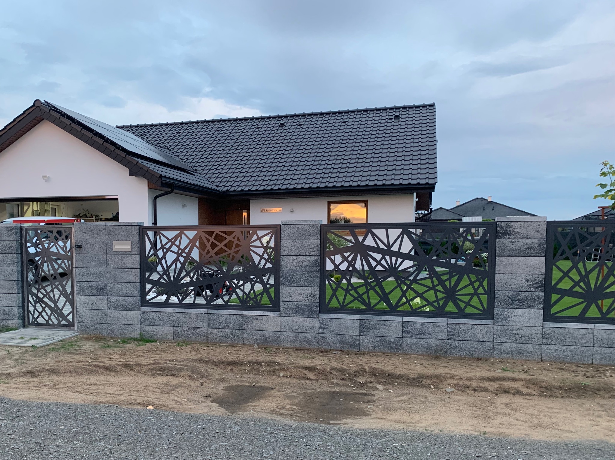 Nowoczesna brama wjazdowa i ogrodzenie panelowe z geometrycznym wzorem, wykonane z metalu i osadzone w szarych, betonowych słupkach, na tle domu z ciemną dachówką i panelami słonecznymi.