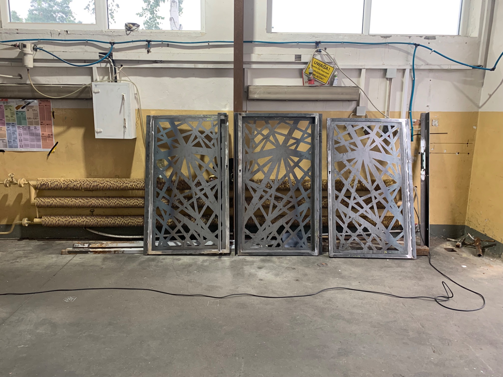 Trzy metalowe panele dekoracyjne z geometrycznym wzorem, ustawione w warsztacie na betonowej podłodze, z grzejnikiem żebrowym i instalacjami widocznymi w tle.
