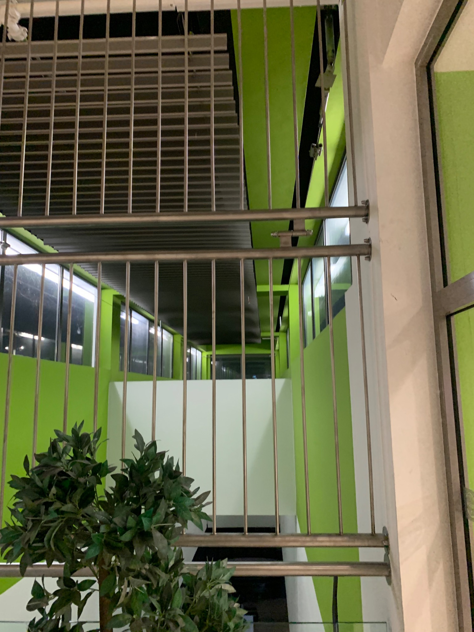 Wewnętrzna balustrada metalowa z pionowymi prętami, widok na korytarz z zielonymi ścianami i białymi elementami, z rośliną doniczkową w dolnej części kadru.