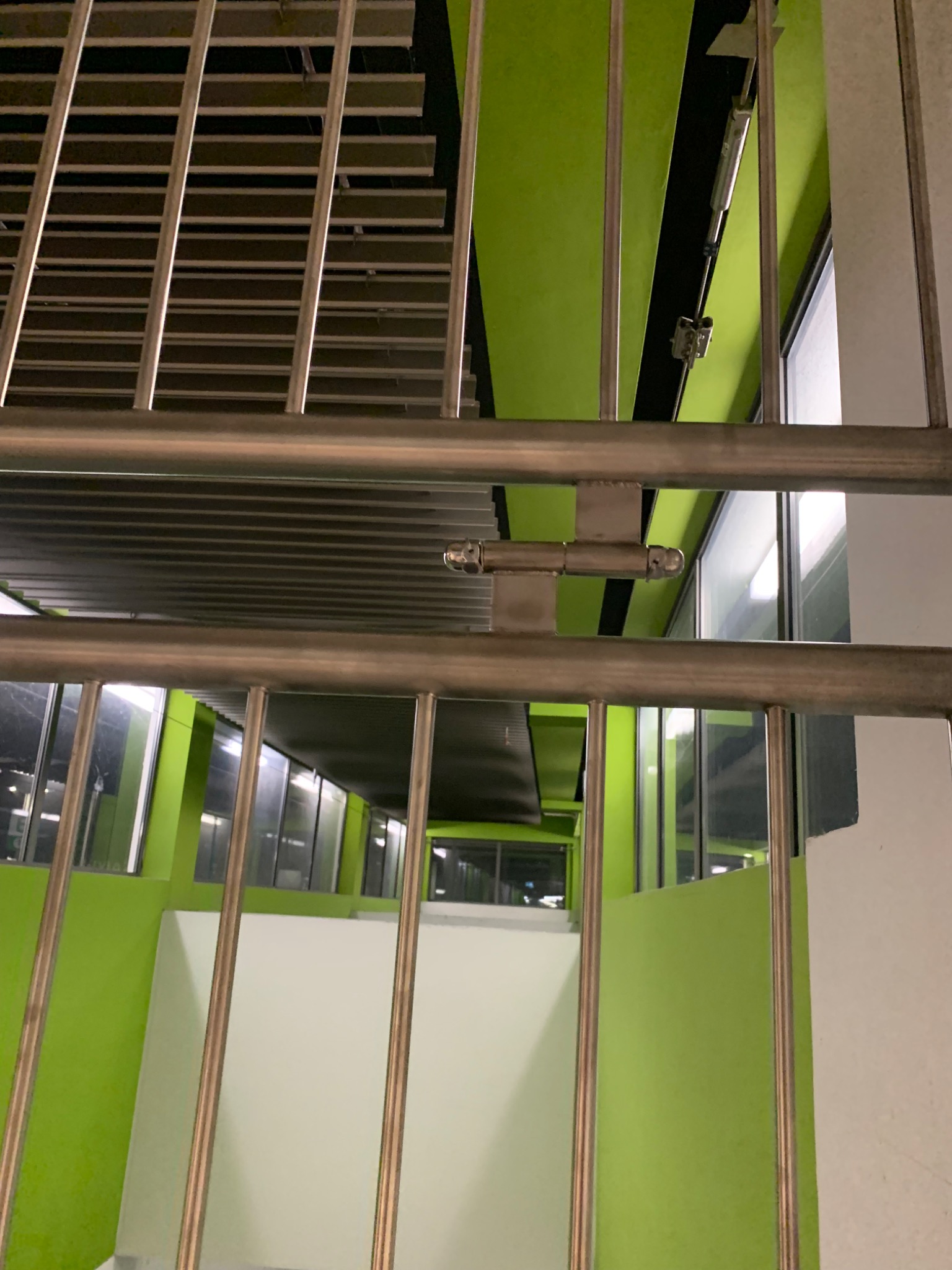 Metalowa barierka z zamkiem zabezpieczającym na tle zielonej ściany i okien w hali przemysłowej.