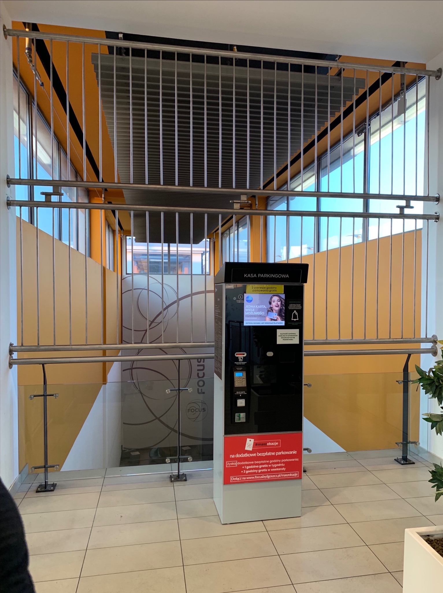 Metalowa konstrukcja balustrady z pionowymi prętami zabezpieczająca schody w centrum handlowym, widoczna kasa parkingowa z reklamą darmowych godzin parkowania.