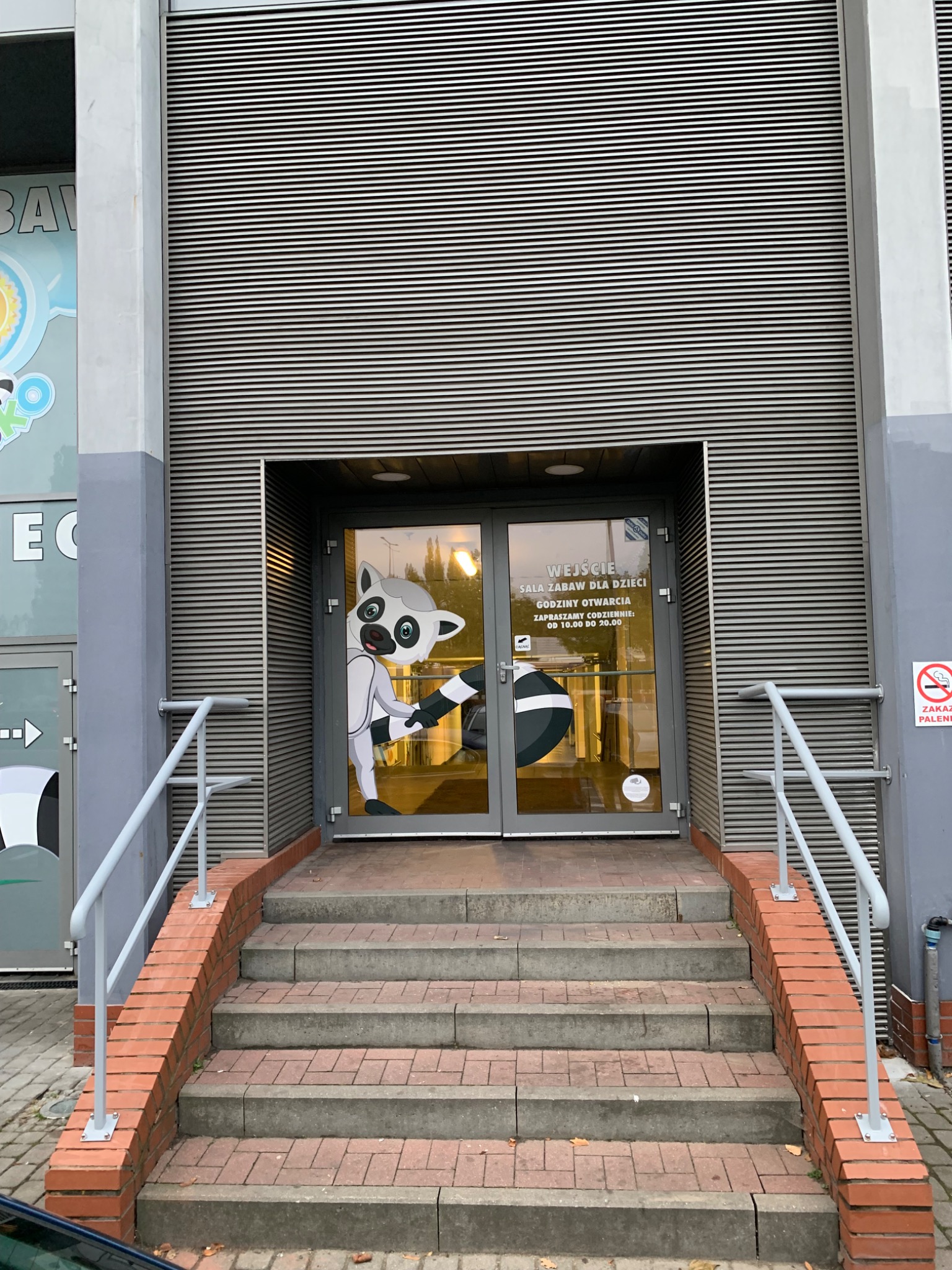 Wejście do sali zabaw dla dzieci, ozdobione naklejką z lemurem, schody z poręczami prowadzą do przeszklonych drzwi.