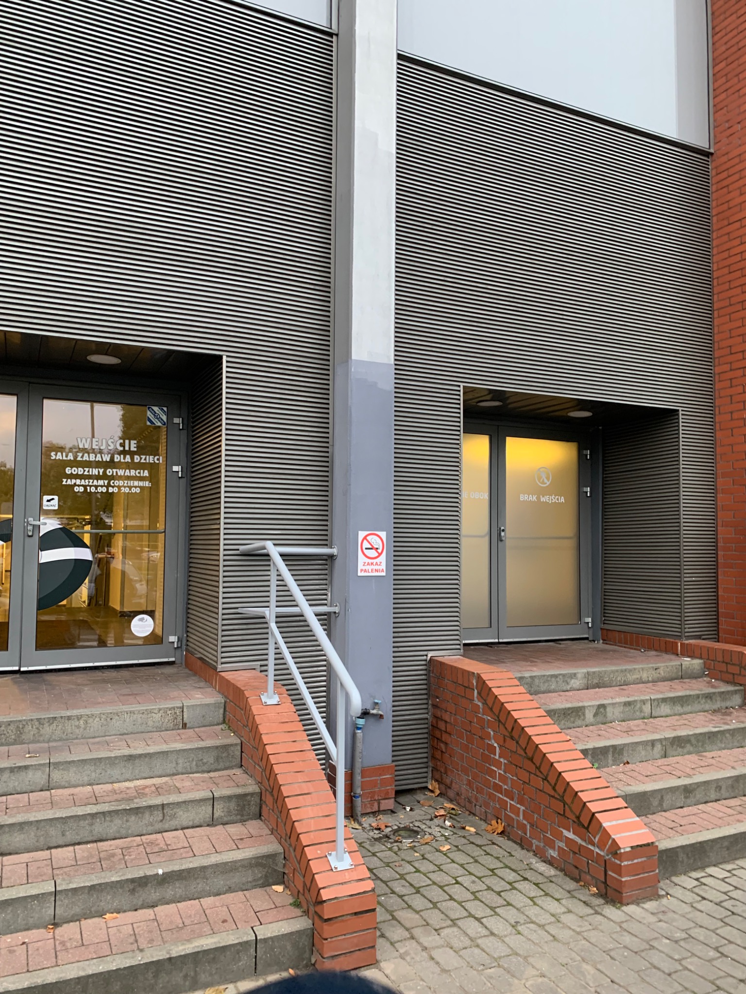 Zewnętrzne schody z ceglanymi bokami i metalową poręczą prowadzące do dwóch wejść w budynku o fasadzie z poziomych, szarych paneli. Na drzwiach po lewej widoczny napis 'Wejście sala zabaw...