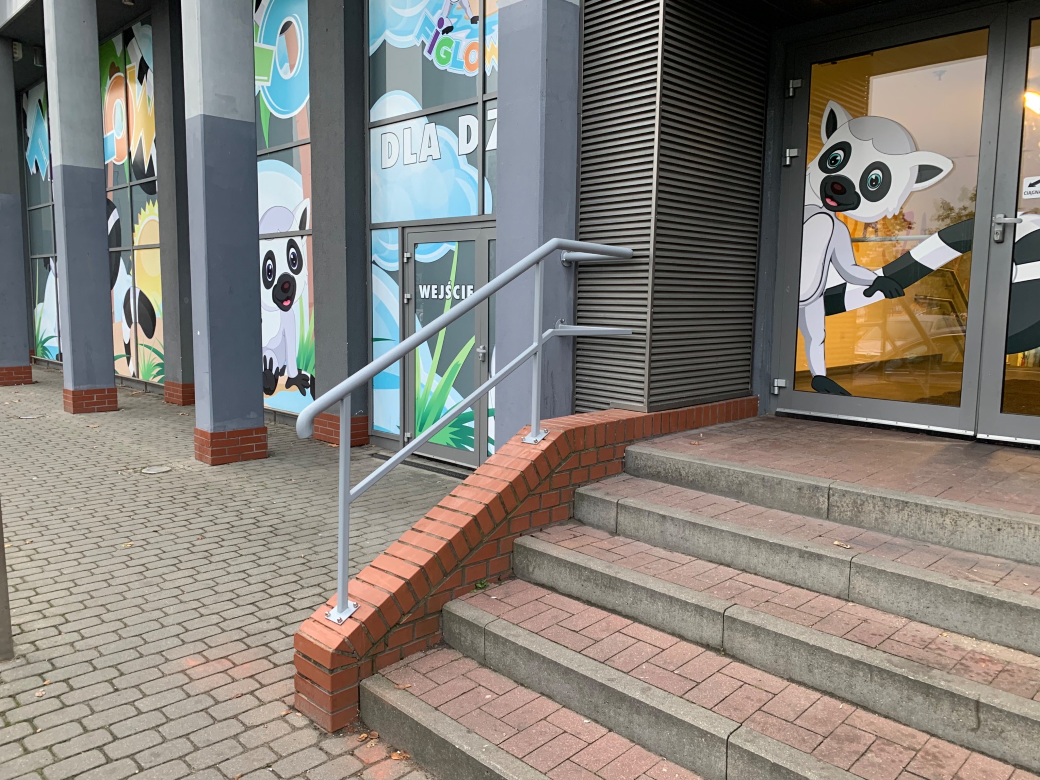 Zewnętrzne schody betonowe z ceglaną obudową i szarą metalową poręczą, prowadzące do wejścia z naklejką lemura.