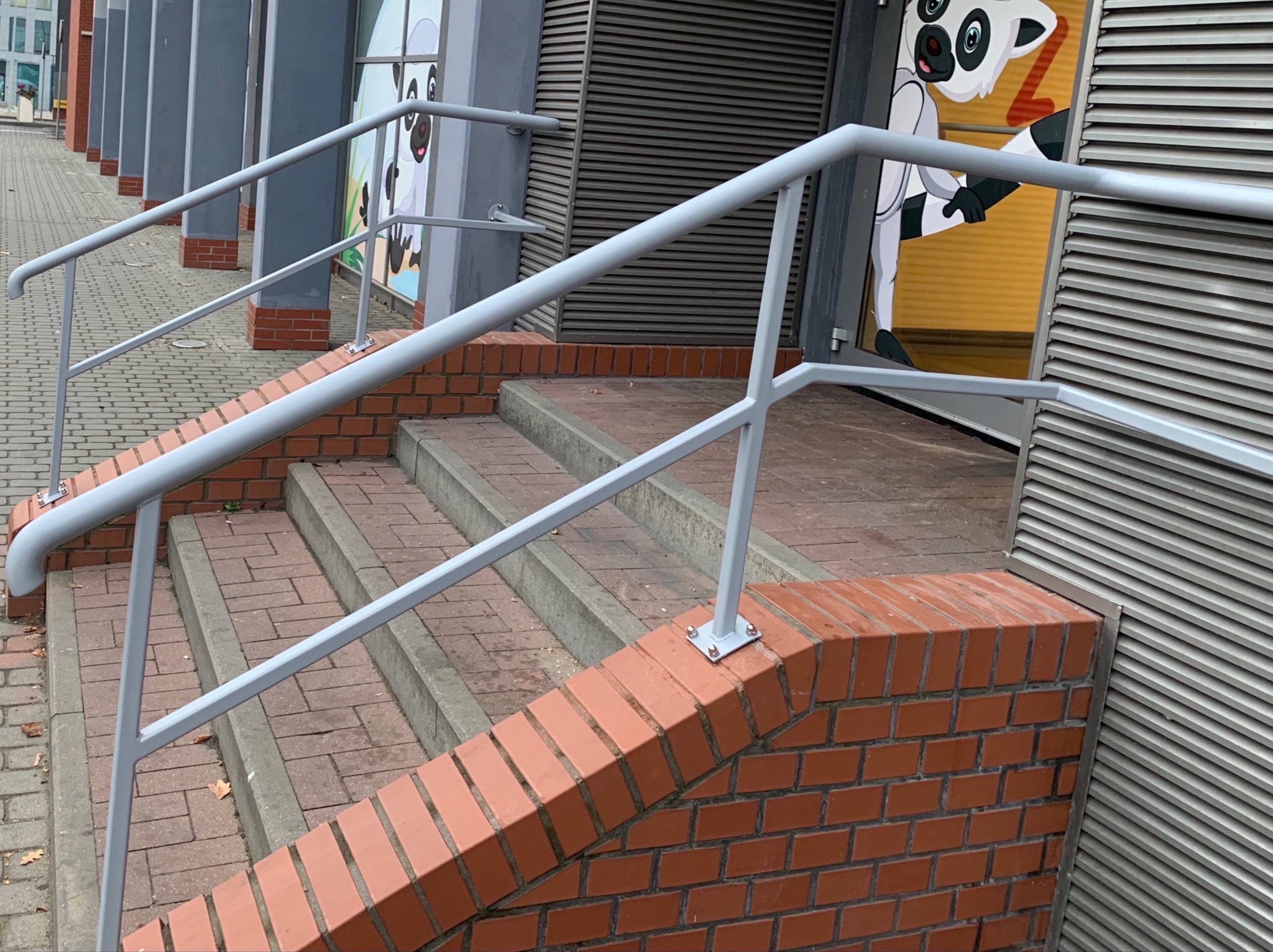 Zewnętrzne schody z ceglanymi obrzeżami i metalowymi poręczami pomalowanymi na szaro, przed budynkiem z szarymi żaluzjami i naklejką z lemurem.