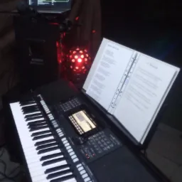 Klawiatura elektroniczna z rozłożonym śpiewnikiem na statywie, obok laptop z oprogramowaniem DJ-skim i mikrofon na tle czerwonej kuli dyskotekowej w ciemnym otoczeniu.