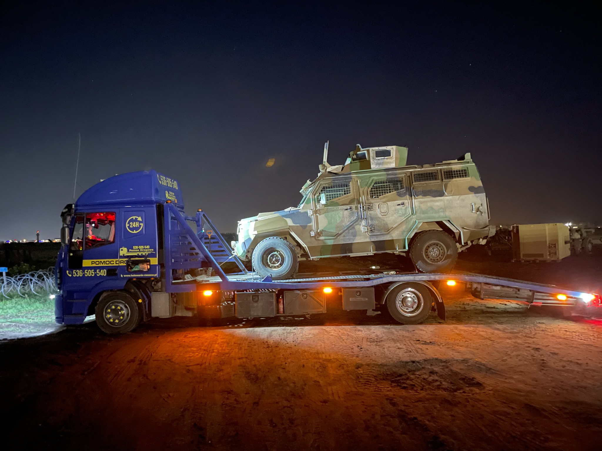 Nocny transport wojskowego pojazdu opancerzonego na niebieskiej lawecie z widocznym numerem telefonu i logiem firmy pomoc drogowa.