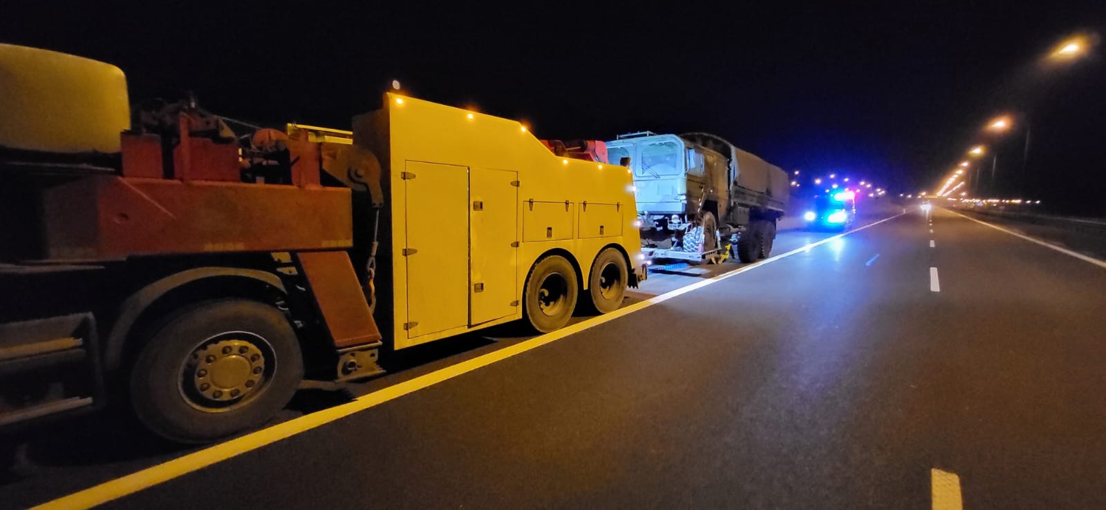 Żółto-czerwona laweta z Rzeszowa holuje wojskowy samochód ciężarowy na autostradzie nocą, w tle światła policyjne.