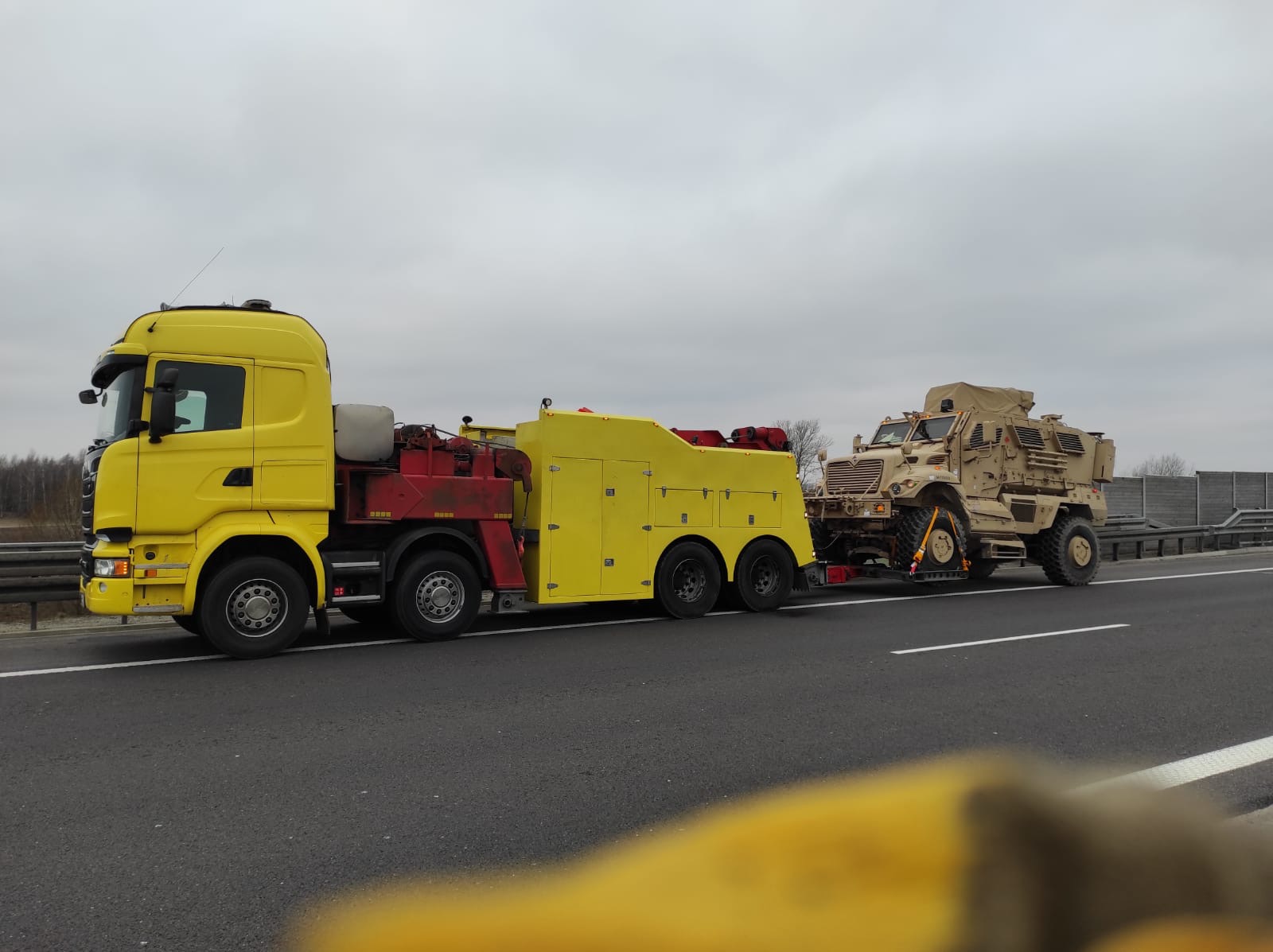 Żółta laweta Scania przewozi wojskowy pojazd opancerzony na autostradzie w pochmurny dzień.