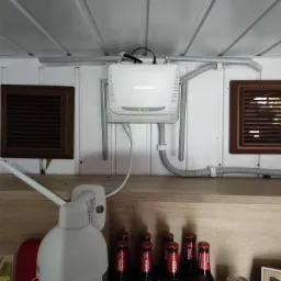 Biały router Mercusys zamontowany na ścianie z drewnianą boazerią, widoczne kable w peszlu, poniżej kamera obrotowa, butelki piwa Lech Free i puszki kawy.