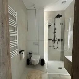 Nowoczesna łazienka w jasnych odcieniach szarości z kabiną prysznicową typu walk-in, białym grzejnikiem drabinkowym i wiszącą toaletą z czarnymi akcesoriami.