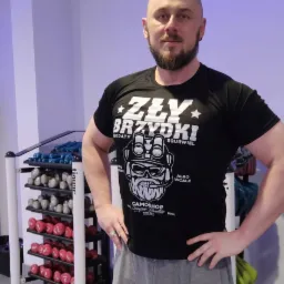 Mężczyzna w czarnej koszulce z nadrukiem, stojący w siłowni na tle regału z hantlami.